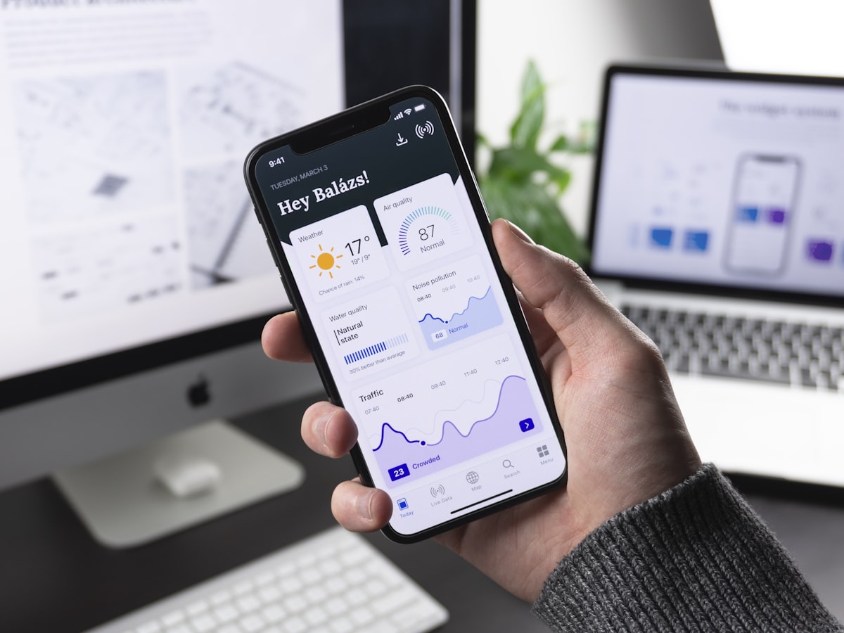 A hand holding a phone showing an app dashboard, a desktop behind
